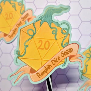 Könnte beinhalten: Eine Reihe von Aufklebern mit einem gelben, kürbisartigen zwanzigseitigen Würfel. Der Würfel hat die Zahl 20 in Orange und ist mit grünen Ranken verziert. Ein Banner mit der Aufschrift "Pumpkin Dice Season" ist um den Würfel gewickelt.