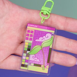 Könnte beinhalten: Ein klarer Acryl-Schlüsselanhänger mit einem grünen Verschluss. Der Schlüsselanhänger zeigt eine Grafik eines Computerfensters mit einer sprießenden Pflanze und dem Text "growth.jpg". Das Design umfasst rosa, grüne und lila Farben.