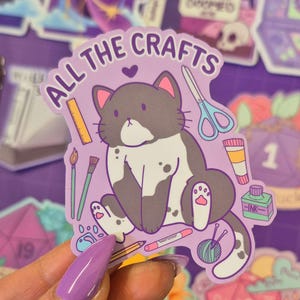 Könnte beinhalten: Ein lila Aufkleber mit einer Cartoon-Katze und dem Text "ALL THE CRAFTS". Die Katze ist grau und weiß mit rosa Akzenten, umgeben von Bastelwerkzeugen wie Schere, Farbe und einem Bleistift. Der Aufkleber wird von einer Person mit lila Nägeln gehalten.
