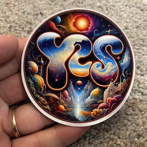 May include: A round sticker with a colorful psychedelic design featuring the word "YES" in a stylized font. The background is a swirling mix of blues, purples, oranges, and yellows, with stars and planets scattered throughout.