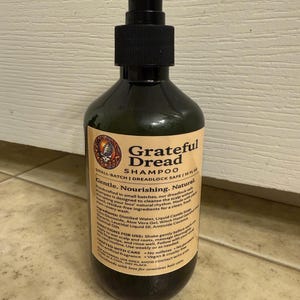 May include: A dark green plastic bottle of "Grateful Dread Shampoo" with a black pump dispenser. The label features text and a circular graphic. The shampoo is described as gentle, nourishing, and natural, designed for dreadlocks. The bottle contains 10 fl oz.