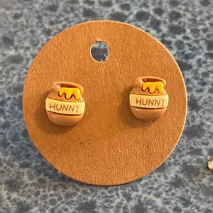 May include: A pair of stud earrings featuring a brown honey pot design with yellow honey dripping from the top. The word "HUNNY" is printed on the front of each pot. The earrings are displayed on a brown circular card.