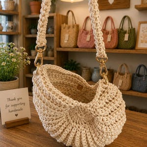 May include: Cream-colored macrame handbag with gold-tone hardware and a braided strap. The bag is displayed on a wooden surface, with a sign that reads "Thank you for supporting handmade." Other handmade bags are visible in the background.