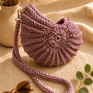 May include: A mauve-colored, handmade crochet bag with a long strap and gold-colored hardware. The bag has a rounded shape with a textured, ribbed design. A pair of brown sunglasses rests near the bag.