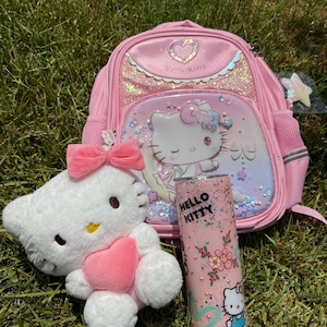 May include: A pink Hello Kitty backpack, tumbler, and plush toy are displayed on grass. The backpack features a heart-shaped design and a Hello Kitty graphic. The tumbler has a floral design and the words "Hello Kitty". The plush toy is white with a pink bow.