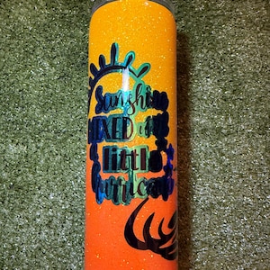 May include: A glittery orange and yellow tumbler with the text "Sunshine Mixed with a Little Hurricane" in black and teal. The tumbler has a clear lid and is cylindrical in shape.