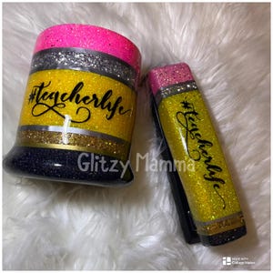 May include: Glittery pencil-themed tumbler and phone grip. The tumbler is yellow with black glitter at the base and pink glitter at the top. The text "#teacherlife" is written on the tumbler. The phone grip is a pencil shape with the same design.