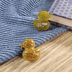 Puede incluir: Dos accesorios dorados con forma de pato con purpurina. Uno es un broche con un cierre plateado, y el otro es una pequeña figura. Están sobre una tela azul y una superficie de madera. Un libro con una cubierta azul y dorada está en el fondo.