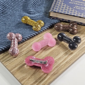 May include: A selection of glittery penis-shaped brooches in gold, brown, pink, and black. One brooch has a pin fastening. The brooches are arranged on a wooden board and a blue cloth.