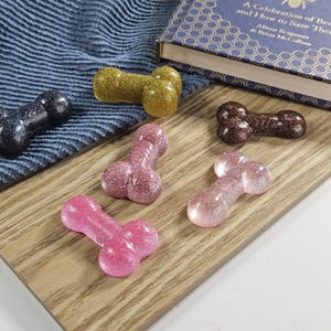Puede incluir: Una variedad de objetos con forma de pene de resina brillante en varios colores, incluyendo rosa, dorado y negro. Los objetos están dispuestos sobre una superficie de madera, con un paño azul y un libro en el fondo.
