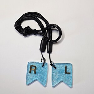 May include: A lanyard featuring two light blue, flag-shaped charms. The charms display gold letters "R" and "L" and are connected to a black cord. The cord has a black clasp and a loop for wearing around the neck.