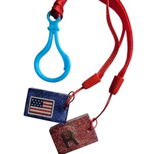 Op de afbeelding: Een rode lanyard met een blauwe plastic sluiting en twee rechthoekige bedels. Eén bedel is blauw met een Amerikaanse vlag, de andere is rood met de zilveren letter "R".