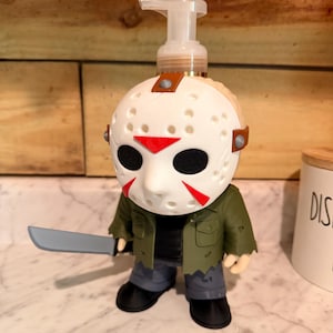 May include: A soap dispenser shaped like a figure wearing a white hockey mask with red triangular markings and black eye holes. The figure wears a green jacket over a black shirt, grey trousers, and black shoes, while holding a grey plastic machete.