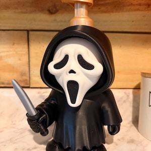 Può includere: Un dispenser di sapone a forma di figura che indossa una tunica nera con cappuccio e una maschera da fantasma bianca. La figura tiene un piccolo coltello grigio nella mano destra. Un meccanismo a pompa in plastica dorata è fissato sul cappuccio.