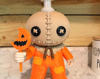 May include: A soap dispenser shaped like a character with a burlap-textured head, black button eyes, and a stitched mouth. The figure wears an orange jumpsuit with yellow patches and holds an orange lollipop with a black jack-o'-lantern face.