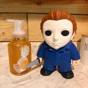 May include: A clear plastic pump bottle filled with amber-colored liquid soap featuring a white pineapple graphic and the text Pineapple Prosecco, positioned next to a stylized figurine with a white mask, brown hair, blue jumpsuit, and a small knife.