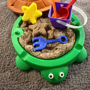May include: A green turtle-shaped sandbox with a brown lid, yellow star, and blue rake. Includes a purple bucket with red and orange tools, and sand. A fun, colorful playset for kids.