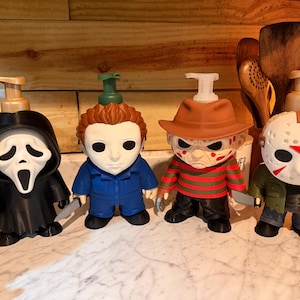 Puede incluir: Cuatro dispensadores de jabón decorativos con forma de personajes de películas de terror. Las figuras incluyen un fantasma con túnica negra, un hombre con mono azul, una figura con sombrero marrón y suéter a rayas, y una máscara de hockey blanca.