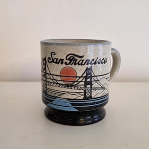 May include: A ceramic mug with a speckled beige finish, featuring a design of the Golden Gate Bridge, a setting sun, and the words "San Francisco" in black script. The mug has a black base and a light gray handle.