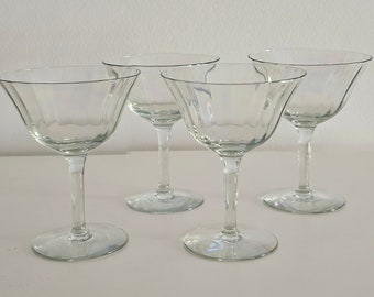 May include: Four clear, stemmed cocktail glasses with a classic coupe design. The glasses have a wide, shallow bowl and a thin stem, perfect for serving cocktails. The glasses are made of clear glass and are arranged on a white surface.
