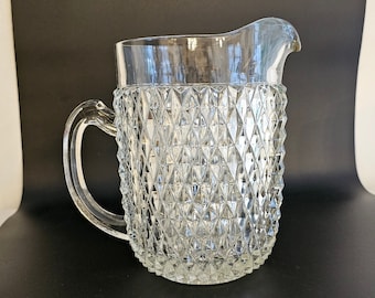 May include: A clear glass pitcher with a diamond pattern on the lower portion and a smooth top. The pitcher has a handle and a spout for pouring. The background is black and white.