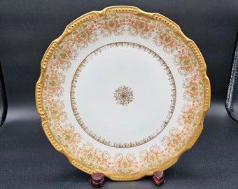 May include: A white porcelain plate with a gold rim and intricate floral pattern in shades of orange, green, and gold. The plate has a scalloped edge and is displayed on a wooden stand.