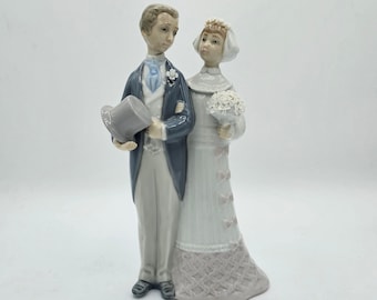 May include: A porcelain figurine of a bride and groom. The bride is wearing a white dress with a floral pattern and a white veil. The groom is wearing a gray suit with a blue tie and a top hat. The figurine is a traditional wedding decoration.