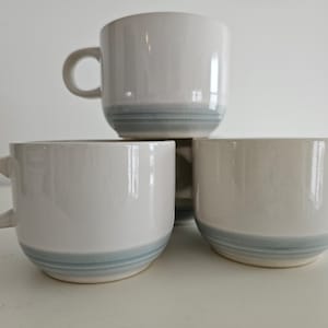 May include: Three white ceramic mugs with a light blue band around the top. The mugs are stacked on top of each other.