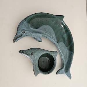 May include: Two-piece ceramic set featuring a dolphin design. The set includes a large, curved dolphin-shaped platter and a smaller dolphin-shaped bowl. The pieces are glazed in a speckled teal color, with a glossy finish.