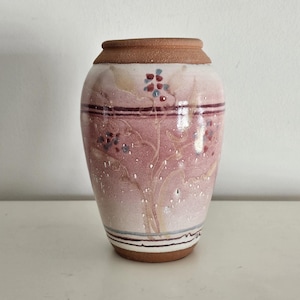 May include: A ceramic vase with a reddish-pink body and a brown rim. The vase features a floral design with red and blue accents. The base has a brown rim with a white surface.