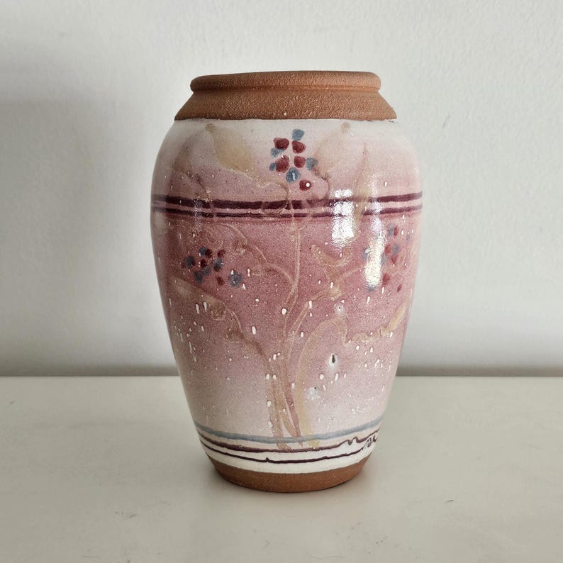 May include: A ceramic vase with a reddish-pink body and a brown rim. The vase features a floral design with red and blue accents. The base has a brown rim with a white surface.