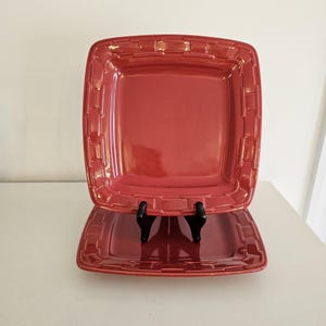 May include: Two square, red ceramic plates with a basketweave design around the edges. The plates are displayed on a white surface, supported by a black stand. The plates have a glossy finish.