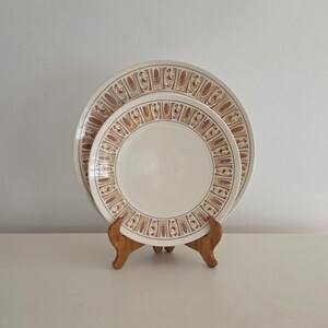 May include: Two round ceramic plates stacked on a wooden stand. The plates are off-white with a wide border featuring a repeating brown and tan pattern of stylized wheat stalks and small bird figures arranged in vertical rectangular panels.