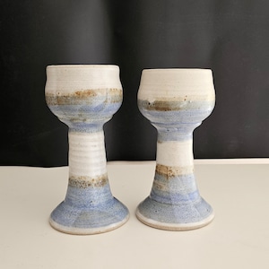 May include: Two handmade ceramic goblets. Each goblet features a white rim, a blue and brown band, and a white stem. The base is a wider blue color. The goblets are displayed on a white surface with a black background.