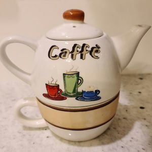 May include: White ceramic teapot with a brown lid and a matching cup. The teapot has the word "Caffe" written on it in brown lettering. The teapot and cup are decorated with three colorful cartoon coffee cups.