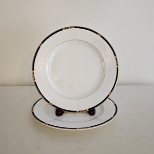 May include: Two white ceramic dinner plates with a black and gold rim and a geometric pattern. The plates are stacked on top of each other.