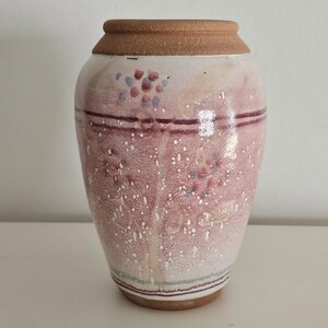 May include: A ceramic vase with a wide base and a narrower neck. The vase is primarily pink with speckled white and small clusters of blue and red dots. Two dark red horizontal lines encircle the vase, and the rim is a light brown color.