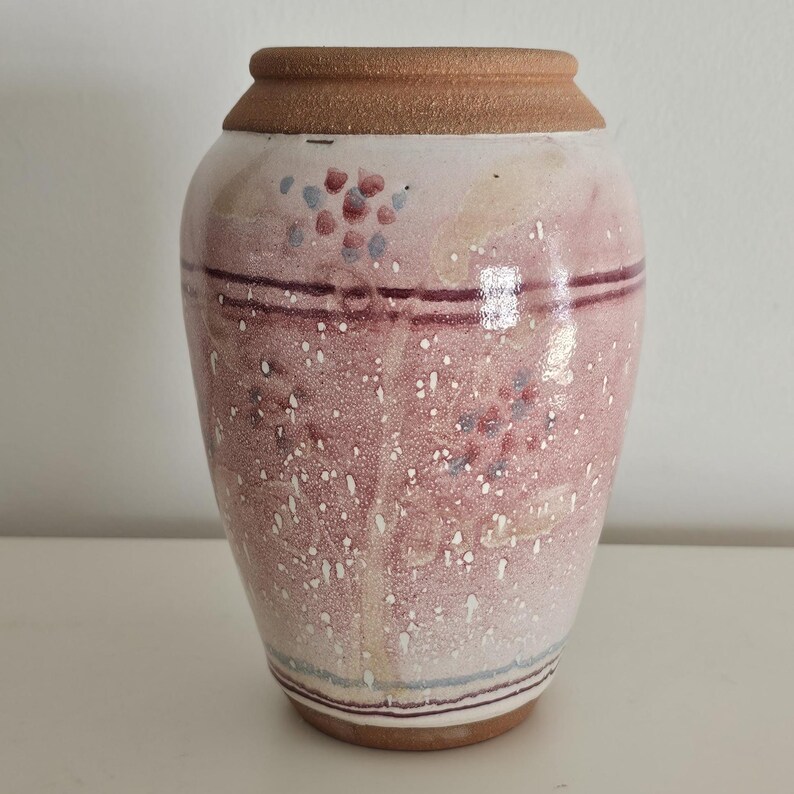 May include: A ceramic vase with a wide base and a narrower neck. The vase is primarily pink with speckled white and small clusters of blue and red dots. Two dark red horizontal lines encircle the vase, and the rim is a light brown color.