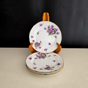 May include: A set of four white porcelain plates featuring a delicate pattern of purple violet flowers and green leaves. Each plate has a scalloped edge with a thin gold trim. The plates are stacked and displayed on a wooden stand.