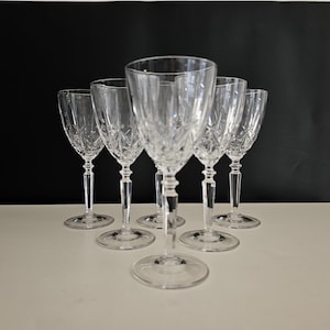 May include: Six clear crystal wine glasses with intricate etched patterns. The glasses have a classic design with a stemmed base and a wide bowl. The glasses are arranged on a white surface against a black background.