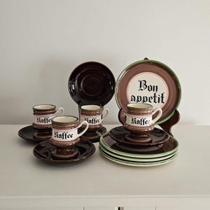 May include: A set of vintage brown and white ceramic coffee cups, saucers, and plates. The cups are labeled "Kaffee" and the large plate says "Bon Appetit". The plates have a green trim. A charming vintage kitchenware set.