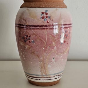 May include: A ceramic vase with a wide base and a narrower top. The vase is primarily pink with a brown rim and base. It features a floral design in red, blue, and tan, with horizontal stripes near the top and bottom.