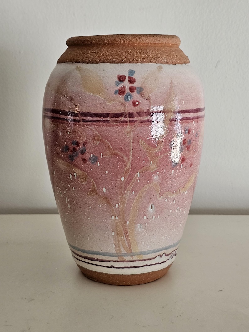 May include: A ceramic vase with a wide base and a narrower top. The vase is primarily pink with a brown rim and base. It features a floral design in red, blue, and tan, with horizontal stripes near the top and bottom.
