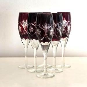 May include: Six ruby red crystal champagne flutes. Each glass has a clear stem and a decorative etched design on the bowl. The etched design includes starburst and leaf patterns. The glasses are arranged on a white surface.