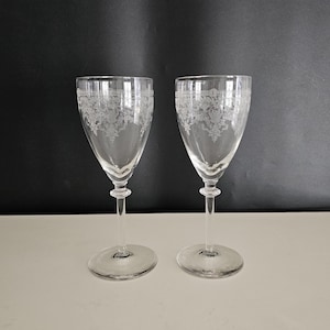 May include: Two clear, stemmed wine glasses with etched floral patterns. The glasses have a rounded bowl, a slender stem with a decorative bulb, and a circular base. The etched design is visible around the top of the bowl.