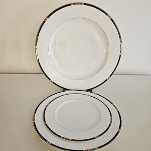 May include: A set of three white ceramic dinner plates with black and gold trim. The plates have a textured surface and a delicate floral design.