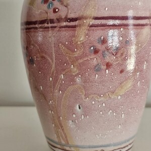 May include: A ceramic vase with a speckled pink and white finish. The vase features hand-painted floral designs in red, blue, and beige. Dark purple and blue bands encircle the vase near the top and bottom, with a brown base.