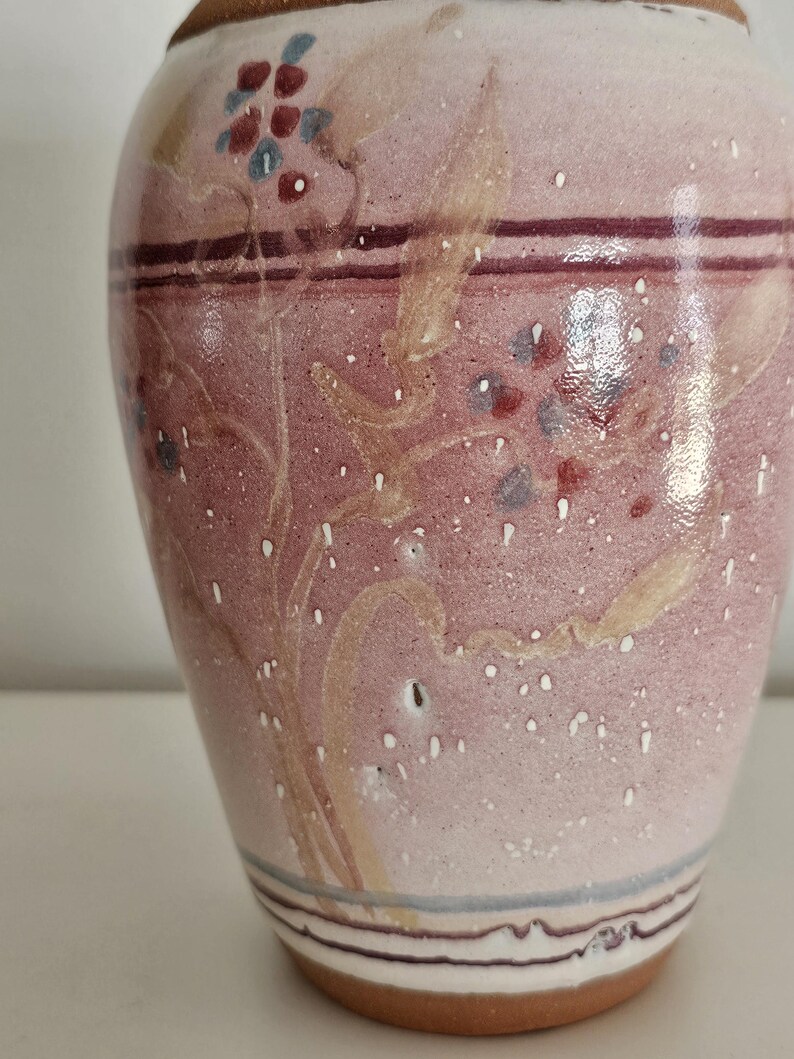 May include: A ceramic vase with a speckled pink and white finish. The vase features hand-painted floral designs in red, blue, and beige. Dark purple and blue bands encircle the vase near the top and bottom, with a brown base.