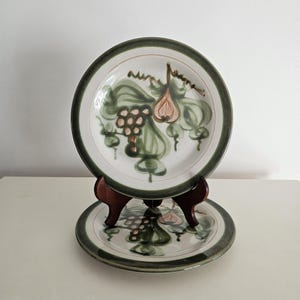 May include: Two ceramic plates with a white background and a green border. Each plate features a hand-painted design of green leaves, grapes, and a peach-colored fruit. The plates are displayed on a wooden stand.