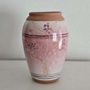 May include: A ceramic vase with a wide base and a narrow neck. The vase is painted with a pink and white floral design, with a brown rim and base. The vase is approximately 7 inches tall.